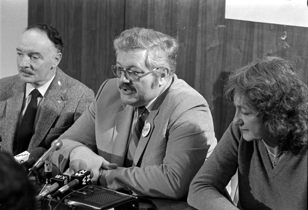 Deux hommes et une femme assis à une table. Quatre micros et un magnétophone à bande sont devant eux. 