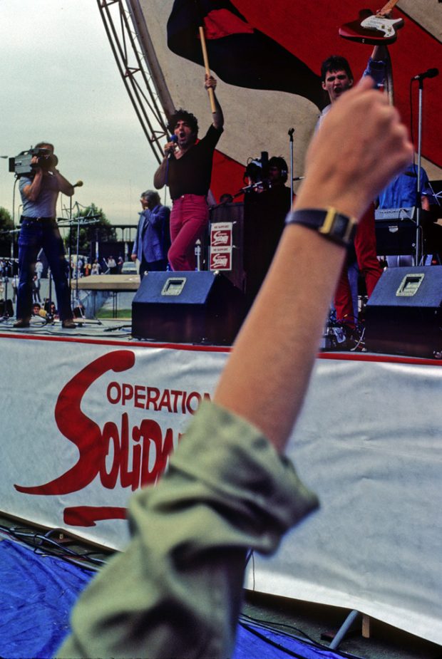 Une scène où des musiciens interprètes sont filmés par un caméraman est ornée du logo d’Operation Solidarity. Au premier plan, on voit le bras levé et le poing serré d’une personne.