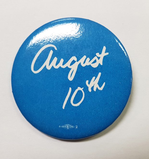 Un macaron bleu et blanc rond présente le script « 10 août ». Sous l’impression, on peut voir un petit logo de syndicat. 