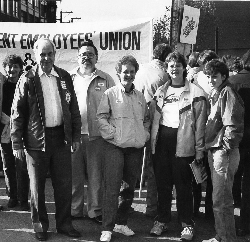 La photo montre cinq personnes portant des macarons et des chandails de Solidarity qui prennent la pose devant une bannière qui indique « Union des employés du gouvernement de la Colombie-Britannique ». On peut voir une pancarte sur laquelle on peut lire « Grève générale! » en arrière-plan. 