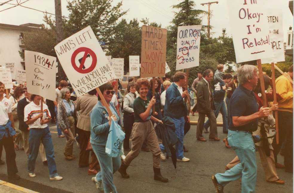 Un groupe de personnes marche dans une parade. Beaucoup tiennent des pancartes qui lisent : « Que reste-t-il lorsque vous n’avez plus de droits? », « Les temps difficiles ne nous arrêteront pas » et « 1984 une année en avance!! »