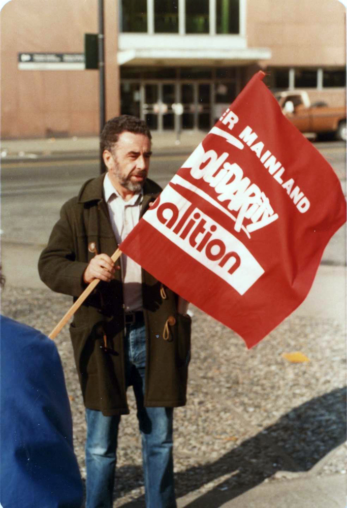 Un homme tiens un drapeau rouge et blanc déroulé sur lequel on peut lire Lower Mainland Solidarity Coalition. 
