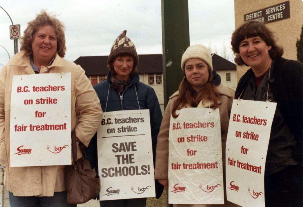 Trois femmes en manteau et chapeau d’hiver se tiennent debout les unes à côté des autres. Elles portent des pancartes sur lesquelles on peut lire « Les enseignants de la C.-B. en grève pour un traitement équitable » et « Les enseignants de la C.-B. en grève, sauvons les écoles! ». Une affiche en arrière-plan indique « Centre de services de district ».