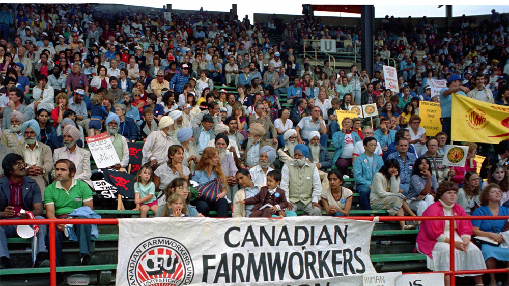 Les personnes et les enfants assis sur les sièges du stade portent des bannières et des affiches. Sur la bannière principale, il est inscrit « Syndicat canadien des travailleurs agricoles ». 