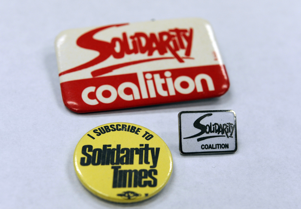 Trois macarons sont affichés, sur lesquels on peut lire : « Solidarity Coalition » en rouge et blanc, « Je m’abonne à Solidarity Times »  en jaune et noir, et une épinglette « Solidarity Coalition » en émail. 
