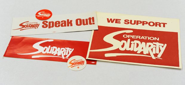 Des autocollants avec le logo d’Operation Solidarity sont montrés. Certains disent « Prenez la parole » et « Nous appuyons ». 