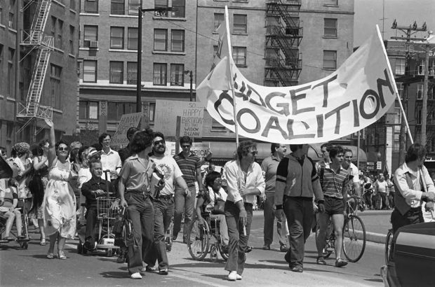 Une parade de gens s’est réunie à pieds, en vélo et en chaises roulantes. Une bannière lit « Budget Coalition » et d’autres pancartes sont visibles. 