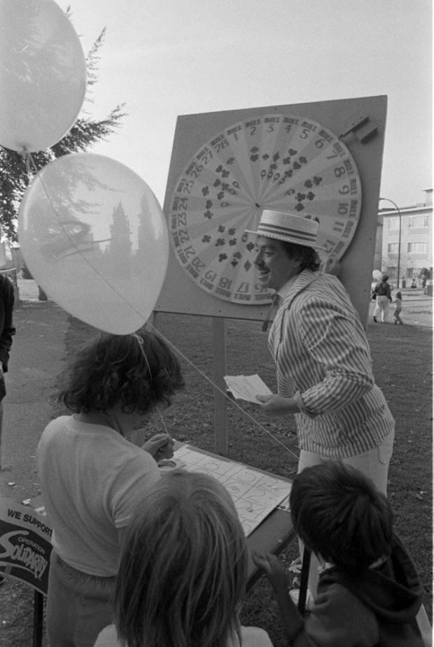 Une photo montre un groupe d’enfants qui regardent un homme tourner une roue de carnaval. Les enfants tiennent des ballons et une pancarte dit « Nous appuyons Operation Solidarity ». 