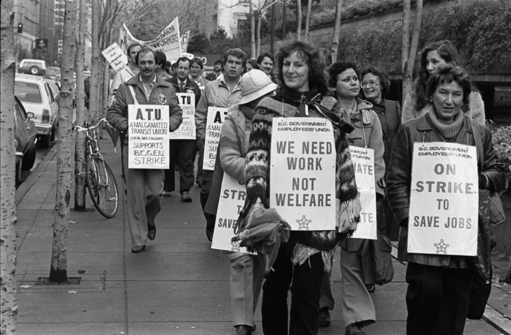 Un groupe de personnes munies de pancartes marchent le long d’un trottoir. Sur les affiches du devant, on peut lire « Union des employés du gouvernement de la C.-B., Nous avons besoin de travail, pas d’aide sociale » et « En grève pour sauver les emplois ». Derrière eux, il y a plus de gens; une affiche indique « ATU — La section locale 583 d’Amalgamated Transit Union de Calgary soutient la grève de la BCGEU ».