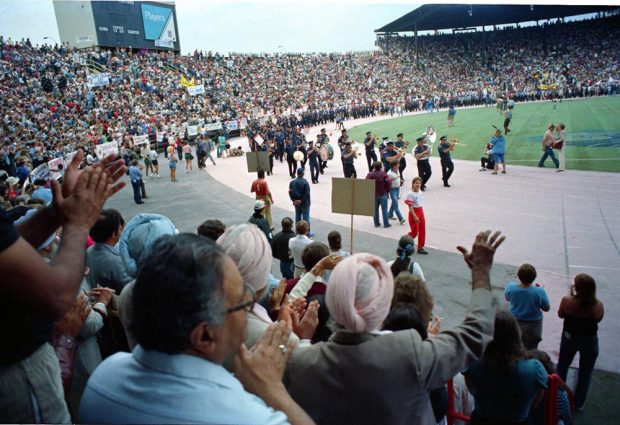 Une photo montre des gens debout qui applaudissent depuis les sièges du stade alors qu’une fanfare dirige une longue parade d’officiers en uniforme. 