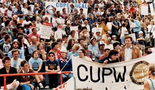 Les sièges du stade sont remplis par les nombreuses personnes présentes et on peut voir les bannières du Syndicat des travailleurs et travailleuses des postes et du Syndicat canadien de la fonction publique. 