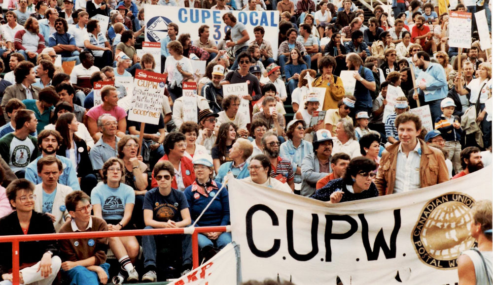 Les sièges du stade sont remplis par les nombreuses personnes présentes et on peut voir les bannières du Syndicat des travailleurs et travailleuses des postes et du Syndicat canadien de la fonction publique. 