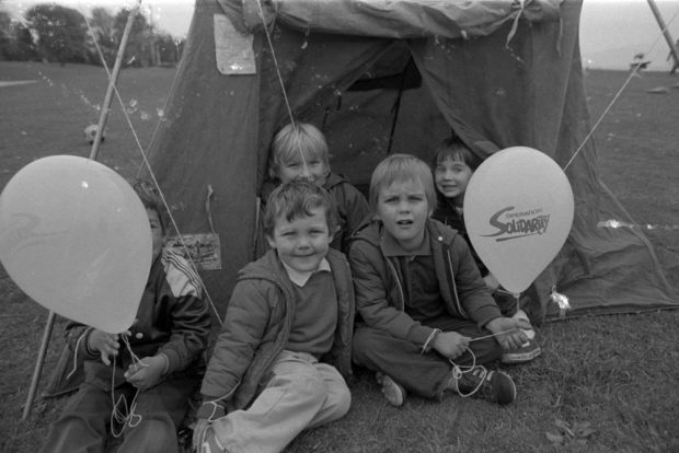 Cinq jeunes enfants sont assis sur l’herbe à l’entrée d’une petite tente. Ils tiennent des ballons affichant le logo d’Operation Solidarity 