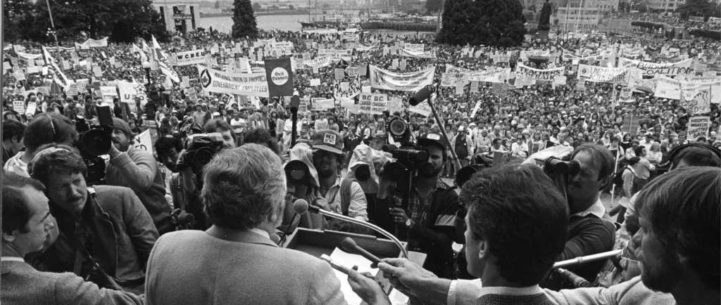 Un homme, prenant la parole devant un podium, fait face à une foule nombreuse sur la pelouse du C.-B. corps législatif. Des journalistes sont rassemblés, tenant des microphones et des caméras vers le podium. Des bannières organisationnelles et des pancartes de protestation sont visibles dans la foule.