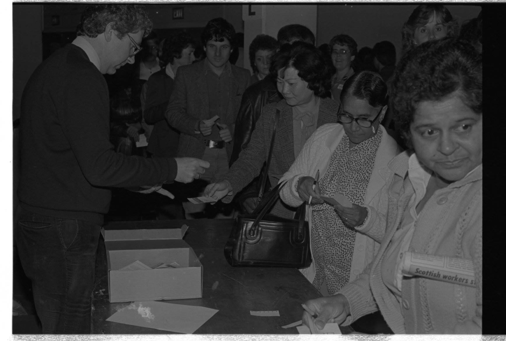 Un homme se tient derrière une table avec une boîte. Des femmes et des hommes font la queue devant la table en tenant des papiers et des stylos. 