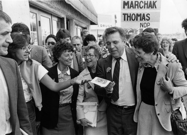 Dave Barrett est entouré par beaucoup de gens souriants alors qu’il marche dans la rue. Il y a Thomas NDP d’écrit sur une pancarte derrière lui. 