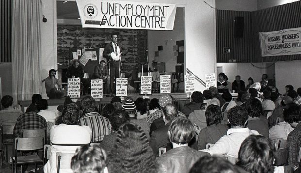 Un homme est au microphone et s’adresse à un public depuis la scène. Trois hommes sont assis sur la scène. On peut lire « Unemployment Action Centre » (Centres d'aide aux chômeurs) sur une banderole au-dessus de la scène et des pancartes sont fixées à la scène.