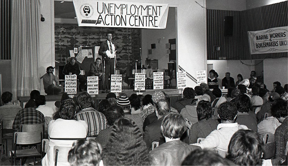 Un homme est au microphone et s’adresse à un public depuis la scène. Trois hommes sont assis sur la scène. On peut lire « Unemployment Action Centre » (Centres d'aide aux chômeurs) sur une banderole au-dessus de la scène et des pancartes sont fixées à la scène.