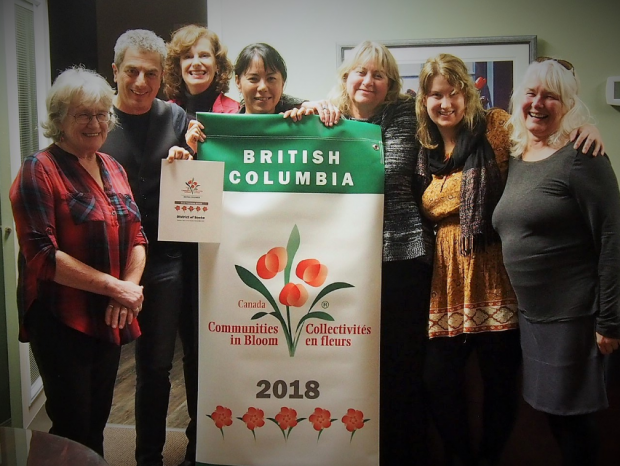 Un groupe de sept personnes debout autour d’une bannière de vinyle que tient une femme, au centre de la photo. Sur cette bannière ornée de motifs floraux, on peut lire : « British Columbia — Canada — Communities in Bloom — Collectivités en Fleurs — 2018 ».