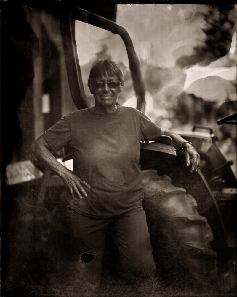Photographie monochrome aux tons sépia d’une femme debout à côté d’un tracteur, le bras reposant sur une portière ouverte.