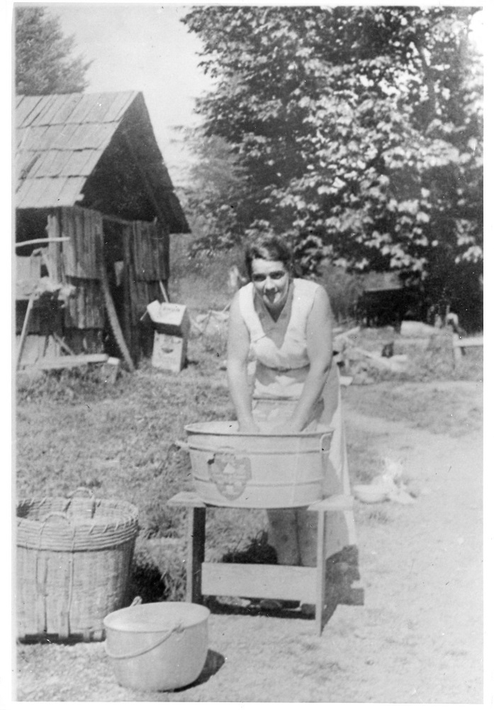 Photographie en noir et blanc d’une femme lavant un vêtement dans une grande cuve à lessive en métal à l’aide d’une planche à laver.
