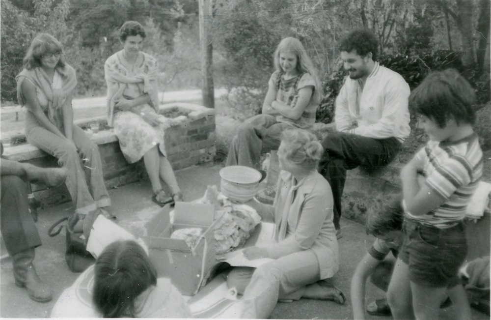 Un groupe d’étudiants assis à l’extérieur, sur le bord de plates-bandes surélevées, autour d’une femme assise au centre.