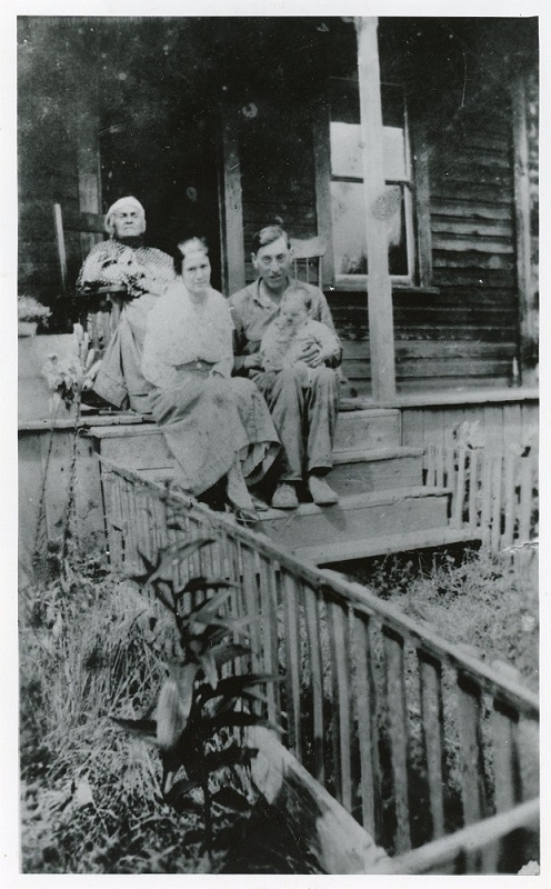 Photographie en noir et blanc d’une femme âgée dans une chaise berceuse en haut des marches d’une véranda; une femme et son mari tenant un bébé sont assis un peu plus bas dans les marches.