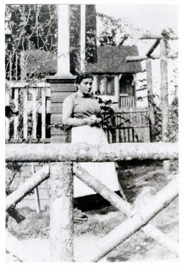 Photographie en noir et blanc d’une femme debout entre un portail de bois et une maison.