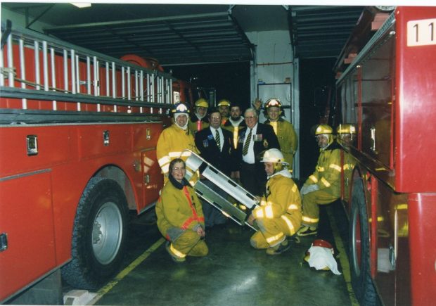 Sept pompiers en tenue de travail et deux hommes en complet portant un insigne, entre deux camions de pompiers.