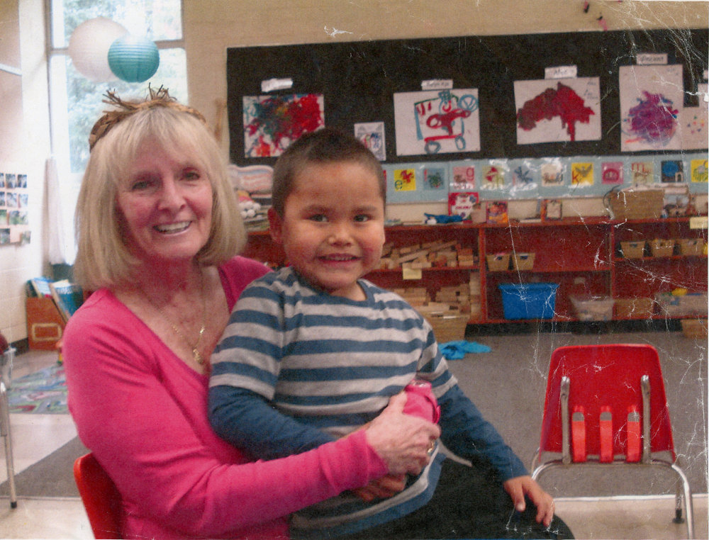 Dans une salle de classe, une femme tenant un petit garçon sur ses genoux. À l’arrière, on voit au mur des peintures faites par les enfants. Il y a aussi des jouets et du matériel d’enseignement dans des paniers.