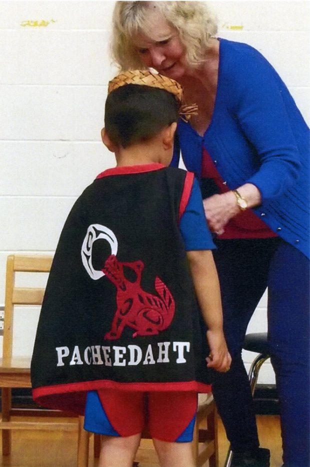 Photo d’une femme ajustant la cape d’un petit garçon. La cape porte le mot « Pacheedaht » et une image d’un loup et de la lune. Le style est caractérisé par les formes et lignes de l’art traditionnel de la côte Nord-Ouest.