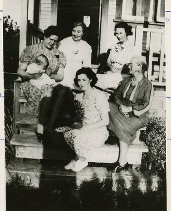 Photographie en noir et blanc de femmes de différentes générations d’une famille, assises ensemble sur des marches de l’entrée d’une maison.