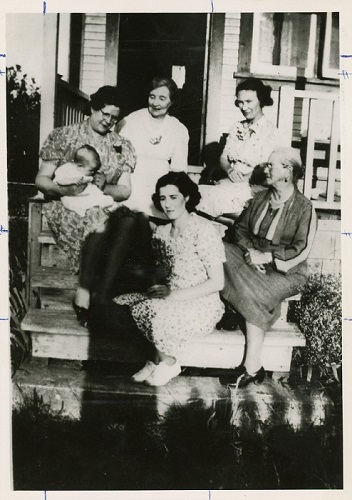 Photographie en noir et blanc de femmes de différentes générations d’une famille, assises ensemble sur des marches de l’entrée d’une maison.