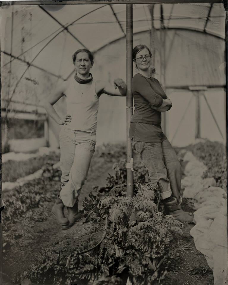 Photographie monochrome aux tons sépia de deux femmes debout s’appuyant sur un poteau, dans une serre.