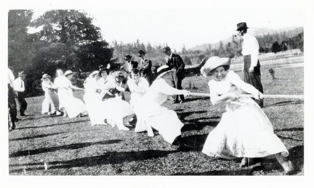 Photographie en noir et blanc de plusieurs femmes souriantes, portant chapeau et robe longue, tirant ensemble de toutes leurs forces sur une longue corde.