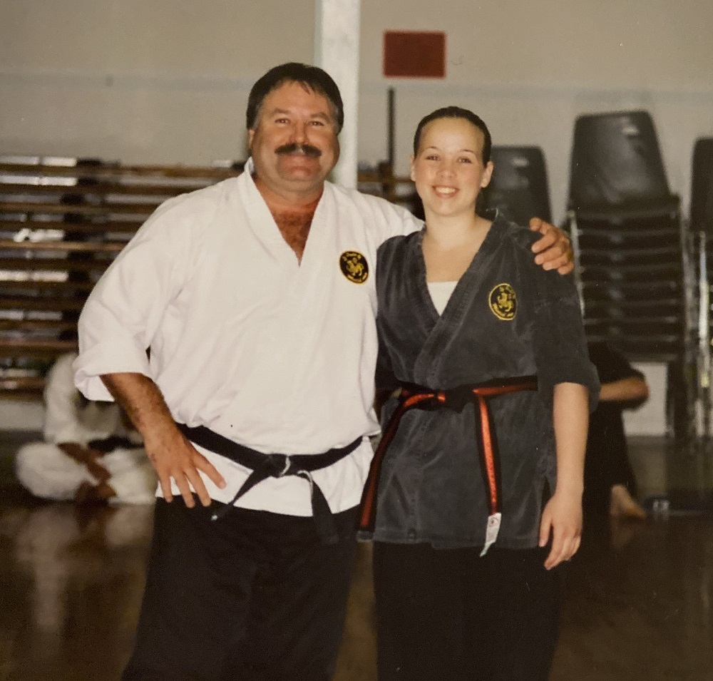 Un homme et une jeune femme sont debout côte à côte, en tenue d’arts martiaux. La ceinture de l’homme est noire et celle de la jeune femme est noire avec un liseré (bande centrale) rouge.