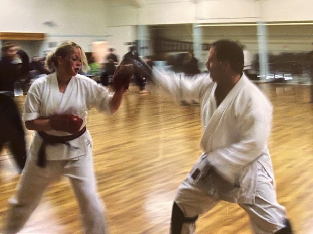Une jeune femme et un homme en tenue d’arts martiaux dans un gymnase, en mouvement, bras levés, en position de frappe et de blocage.