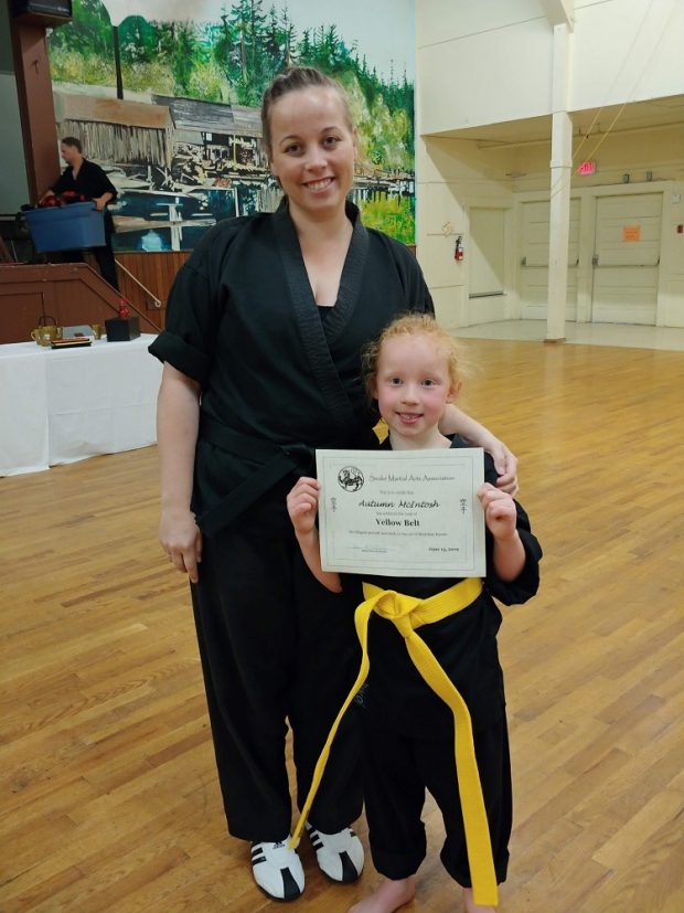 Une femme, la main sur l’épaule de sa fille qui, elle, tient un certificat de promotion à la ceinture jaune.