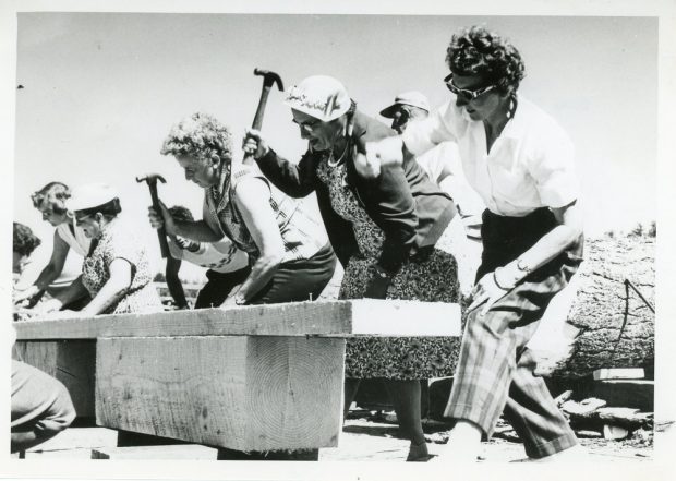 Photographie en noir et blanc d’une rangée de femmes, marteau en main, bras levé, enfonçant des clous dans un morceau de bois.