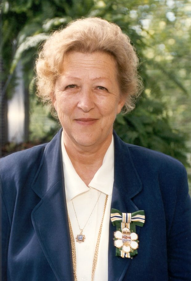 Portrait photo d’une femme portant à son revers un insigne représentant une fleur de cornouiller stylisée sous une boucle de ruban vert, or, blanc et bleu.