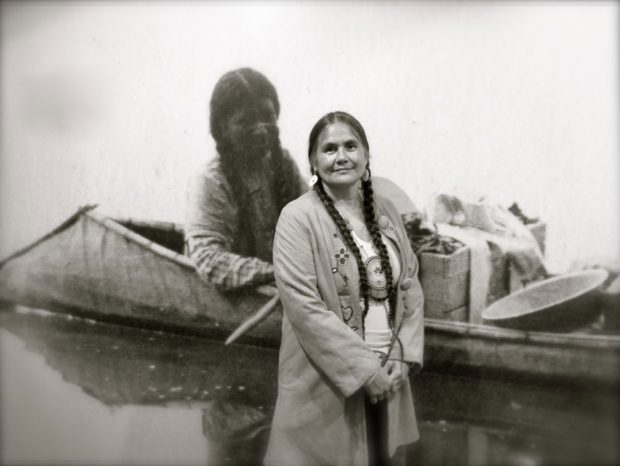 Cette photographie en noir et blanc montre une femme autochtone devant une photographie d’archives agrandie de femmes autochtones dans un canot dont la poupe s’incline vers l’eau. Les deux femmes ont de longs cheveux noirs tressés.