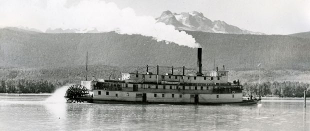 Un bateau à trois étages propulsé par une roue à aubes actionné à la vapeur se déplace sur l’eau. À l’arrière-plan, on aperçoit le rivage et les sommets des montagnes.