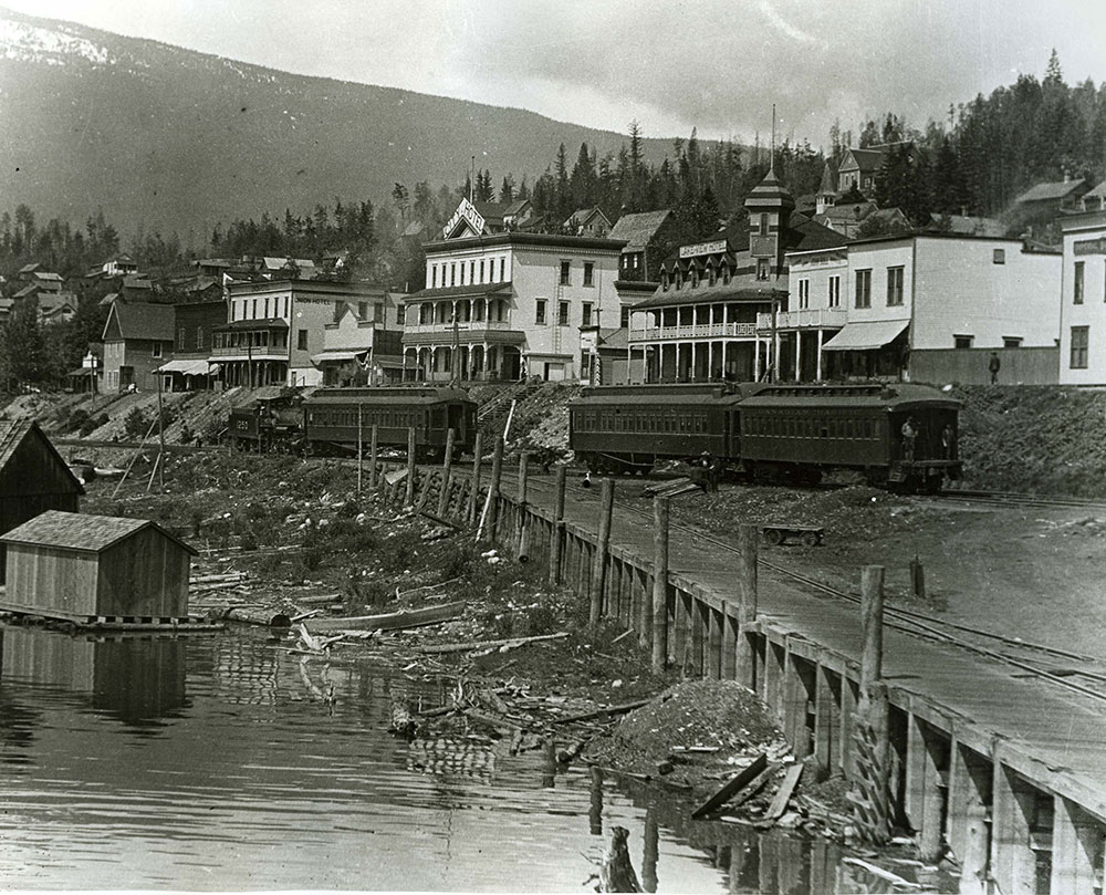 This black-and-white photograph captures a town by the water. The street has store buildings and homes. In front of the buildings is a rail line with train cars on the track. A dock is in front of the train tracks. Part of a mountain and trees are in the background.