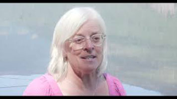 Eileen Pearkes – une femme blanche aux cheveux blancs. Elle porte des lunettes blanches, un collier argenté et une chemise rose. Elle se trouve devant une rivière.