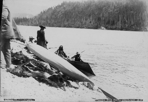 Photographie en noir et blanc montrant deux hommes autochtones à bord d’un canot sur l’eau. Des personnes habillées à l’européenne se tiennent sur la rive des lacs Arrow. Des peaux sont empilées sous un autre canot juste à côté, sur la rive.