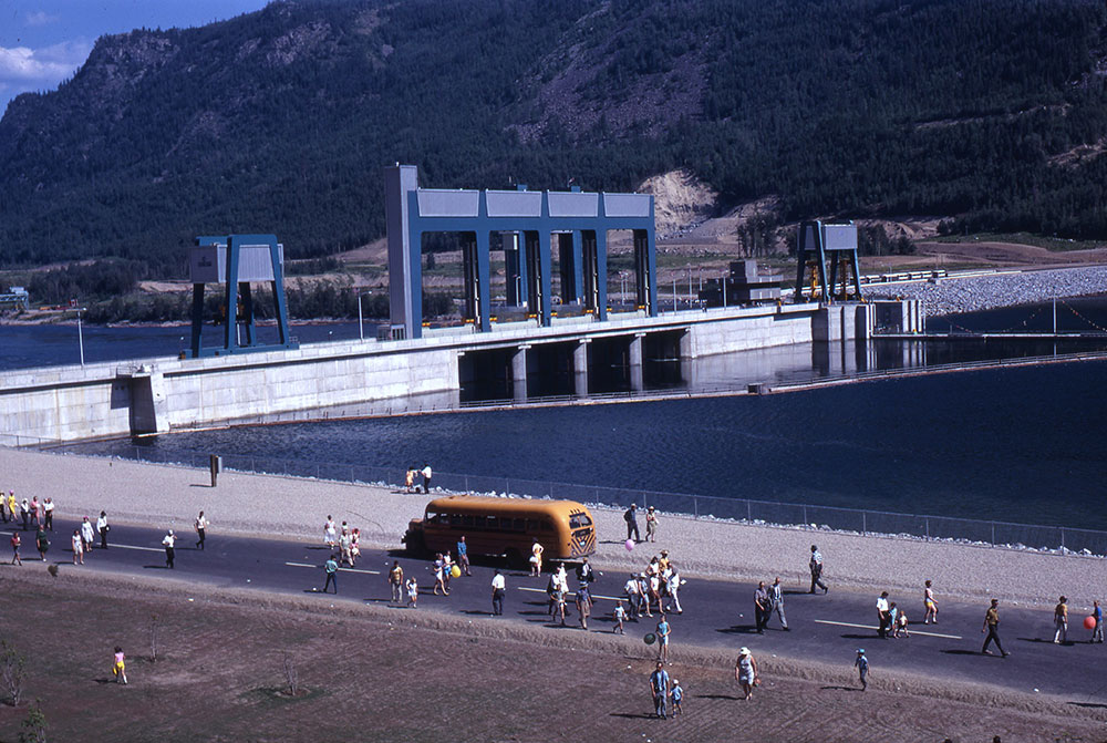 Sur une route, une foule d’environ une cinquantaine de personnes et un autobus se déplacent vers le barrage qui se trouve en arrière-plan. Une partie d'une montagne y est visible derrière.