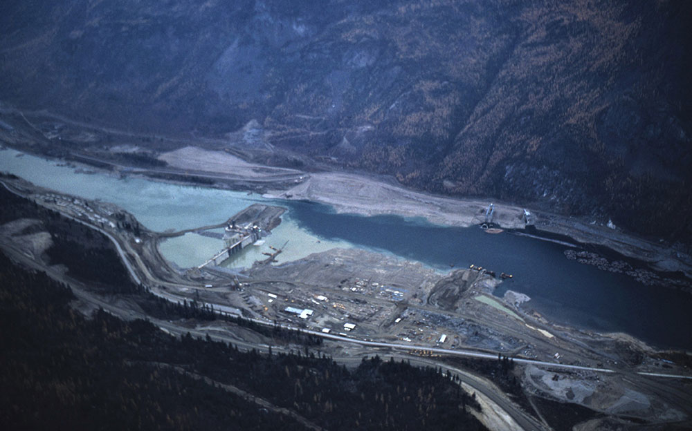 Photographie panoramique plongeante du barrage en construction. On peut y repérer dans l'image le chantier de construction, le barrage, l'eau et les montagnes de l'autre côté.