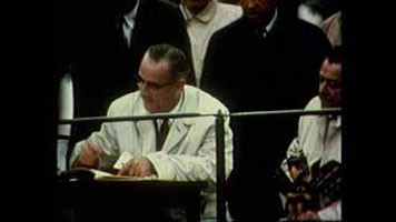 Photo d'un homme assis, vêtu d'un veston de couleur crème signant un document. D'autres personnes sont debout autour de lui en arrière-plan.