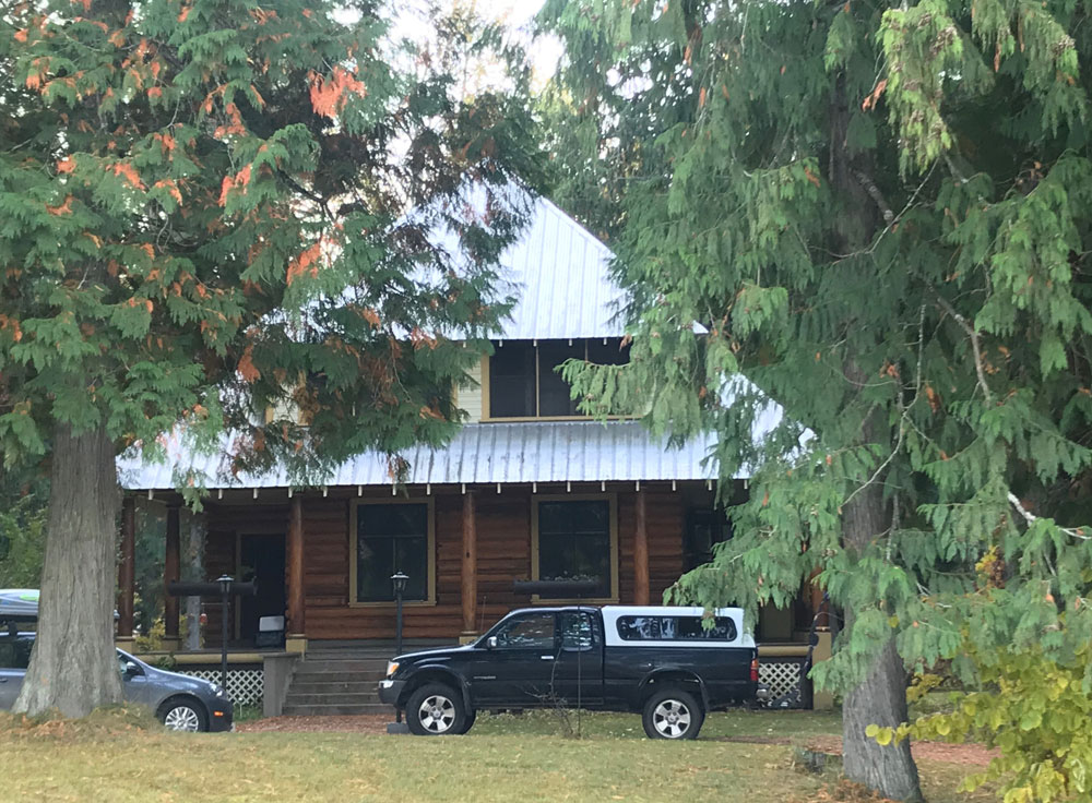 Photographie couleur d'une maison en bois rond à deux étages voilée par deux grands arbres devant. Une voiture et un camion se trouvent devant la maison.
