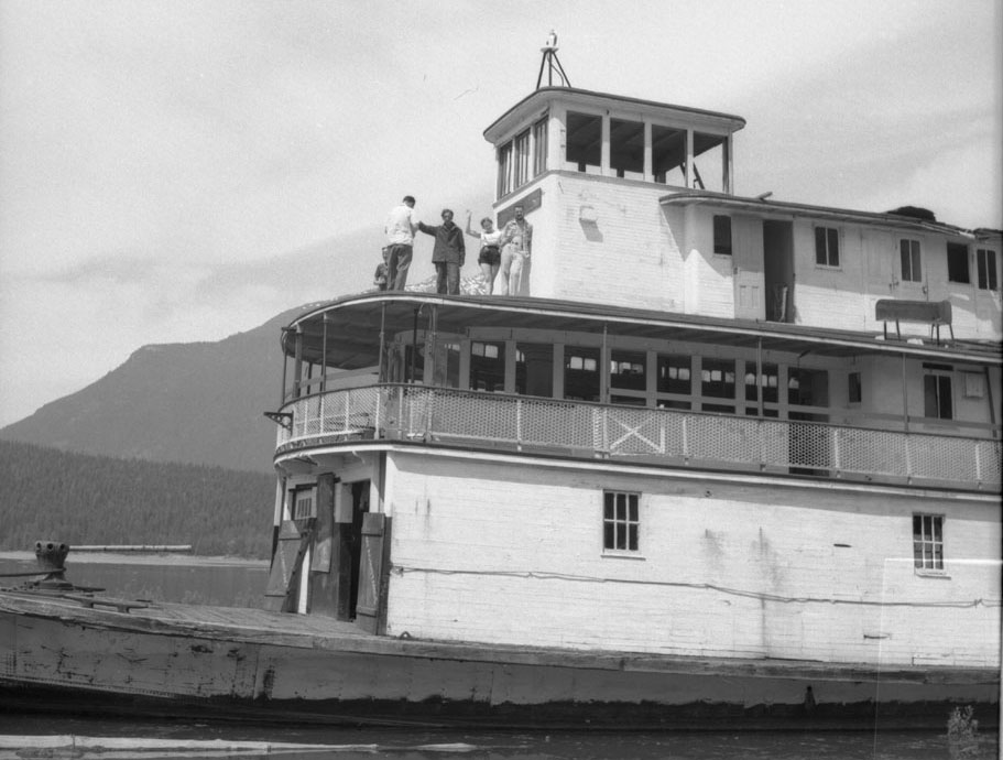 Photographie en noir et blanc de quatre personnes debout sur le troisième étage d’un bateau à vapeur. Seulement le devant du bateau est visible dans la photo. Il y a une montagne et de l’eau à l’arrière-plan, sur le côté gauche.
